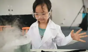 Niña con bata y gafas realiza un experimento científico para niños, creando humo blanco en un cubo metálico.