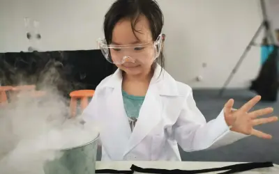 Niña con bata y gafas realiza un experimento científico para niños, creando humo blanco en un cubo metálico.