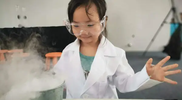Niña con bata y gafas realiza un experimento científico para niños, creando humo blanco en un cubo metálico.