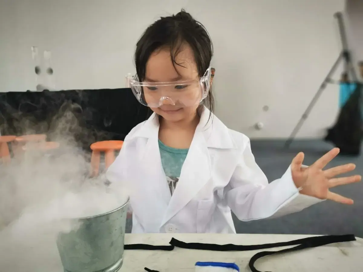 Niña con bata y gafas realiza un experimento científico para niños, creando humo blanco en un cubo metálico.