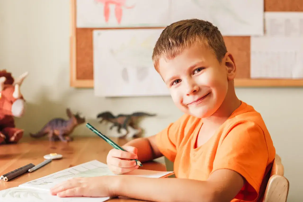 Ni&ntilde;o sonriente practica escritura manual con un l&aacute;piz verde, rodeado de dinosaurios de juguete y dibujos.