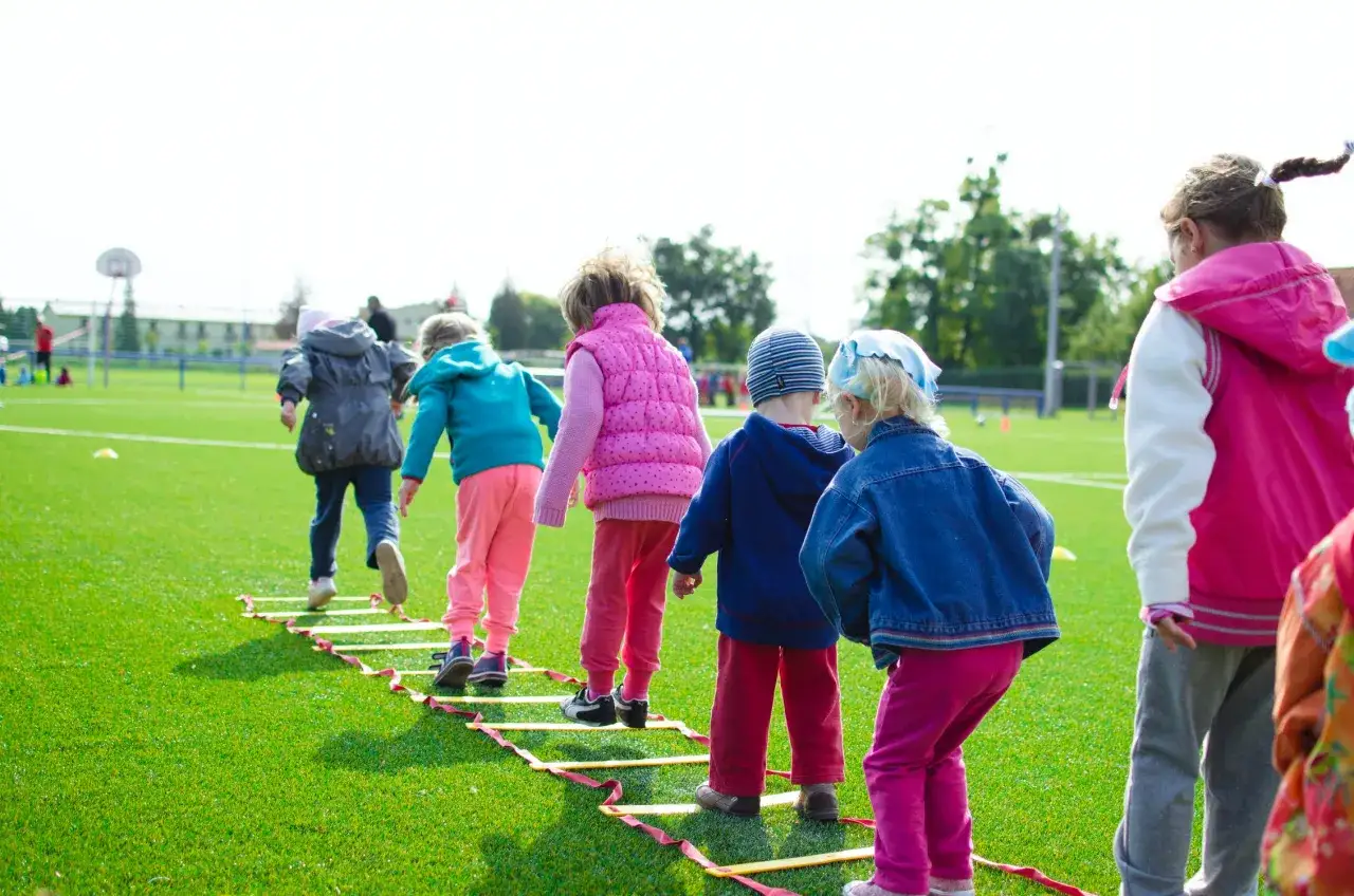 Niños participan en actividades infantiles, corriendo en una escalera de agilidad en un campo de césped artificial.
