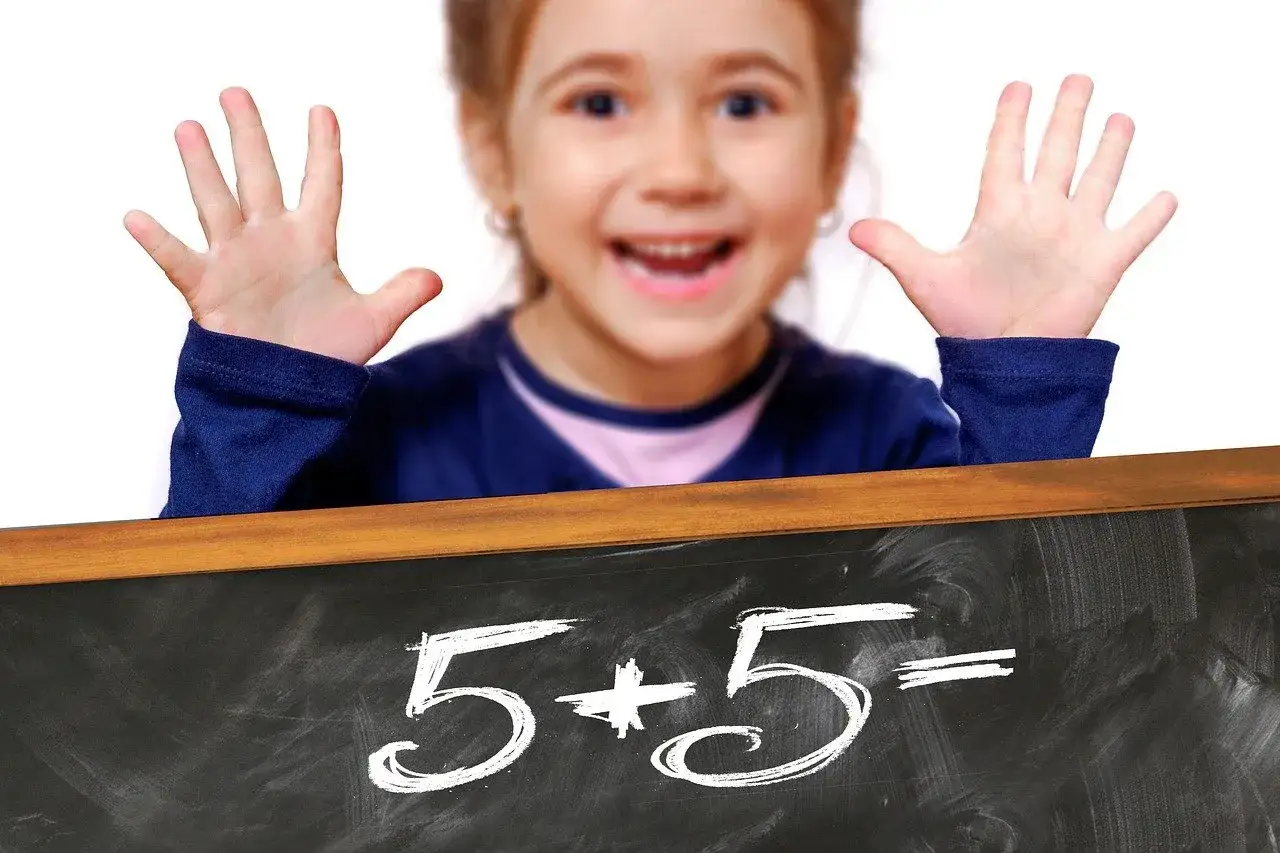 Ni&ntilde;a feliz muestra sus manos, lista para el repaso de matem&aacute;ticas de 2 primaria. En la pizarra, 5+5=.
