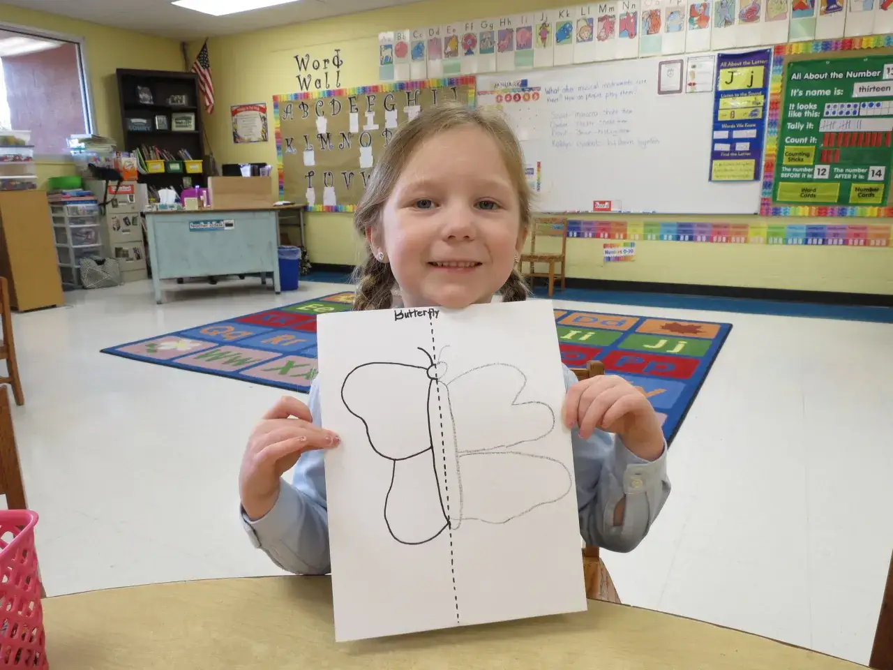 Niña sonriente muestra dibujo de mariposa, aprendiendo sobre simetría primaria en clase.