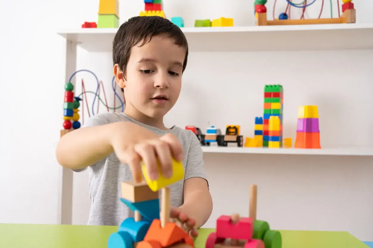 Niño concentrado construyendo con bloques de madera, ideales para actividades para niños de 3 años. Juguetes coloridos en estanterías.