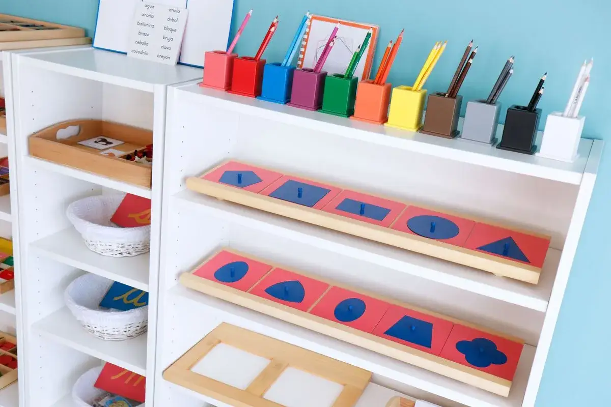 Estanter&iacute;as con materiales educativos que muestran qu&eacute; es Montessori: encajables de formas geom&eacute;tricas, cestas con tarjetas y portal&aacute;pices de colores.