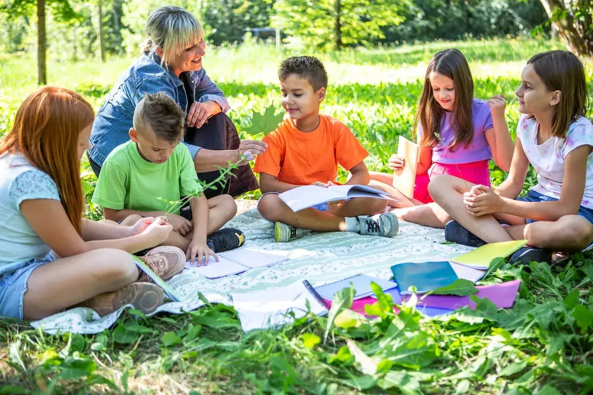 Maestra gu&iacute;a a ni&ntilde;os en juegos de conocimiento del medio en un d&iacute;a soleado. Aprenden sobre la naturaleza mientras est&aacute;n sentados en el c&eacute;sped con cuadernos.