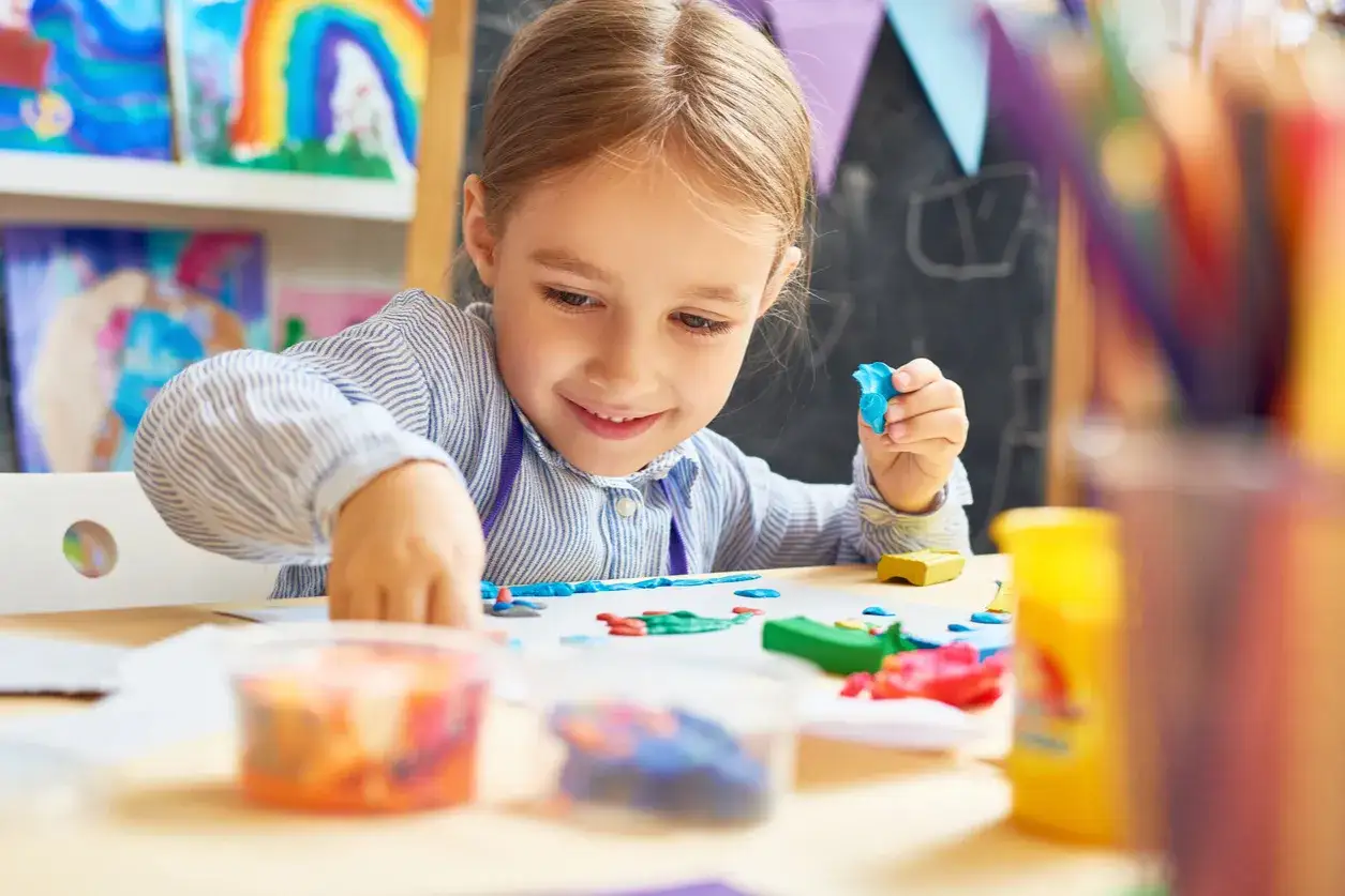 Niña de 0-3 años sonríe mientras moldea plastilina azul. En la mesa hay más plastilina de colores y dibujos.