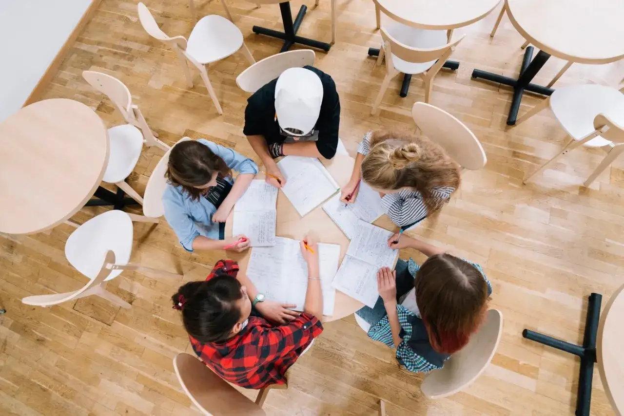 Cinco jóvenes participan en dinámicas de grupo, sentados alrededor de una mesa redonda, escribiendo y colaborando en sus estudios.