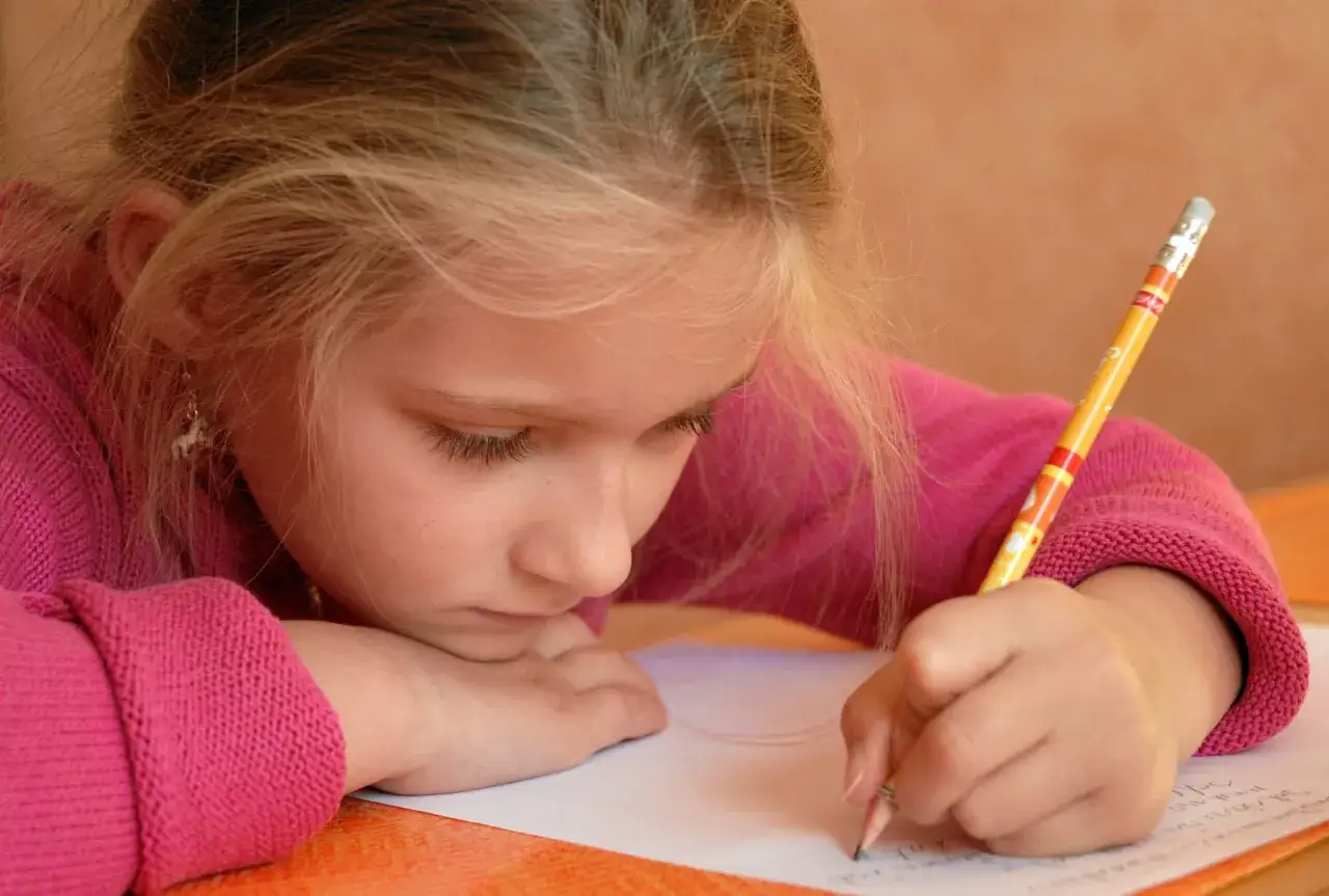 Niña rubia con suéter rosa practica cómo mejorar la letra de un niño, concentrada en su cuaderno.