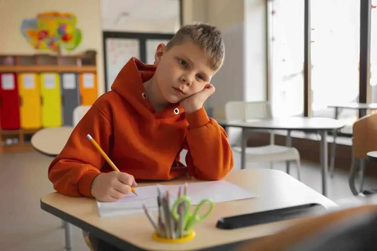 Un ni&ntilde;o con expresi&oacute;n triste en clase, apoyado en su mano, intentando escribir. Una se&ntilde;al de c&oacute;mo detectar el bullying.